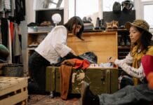 Two women sorting through vintage clothing in a thrift shop, surrounded by shoes and hats.