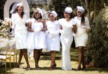 Five women in elegant white outfits enjoying a garden party on a sunny day.