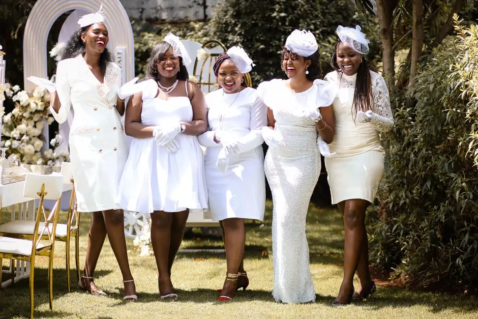Five women in elegant white outfits enjoying a garden party on a sunny day.